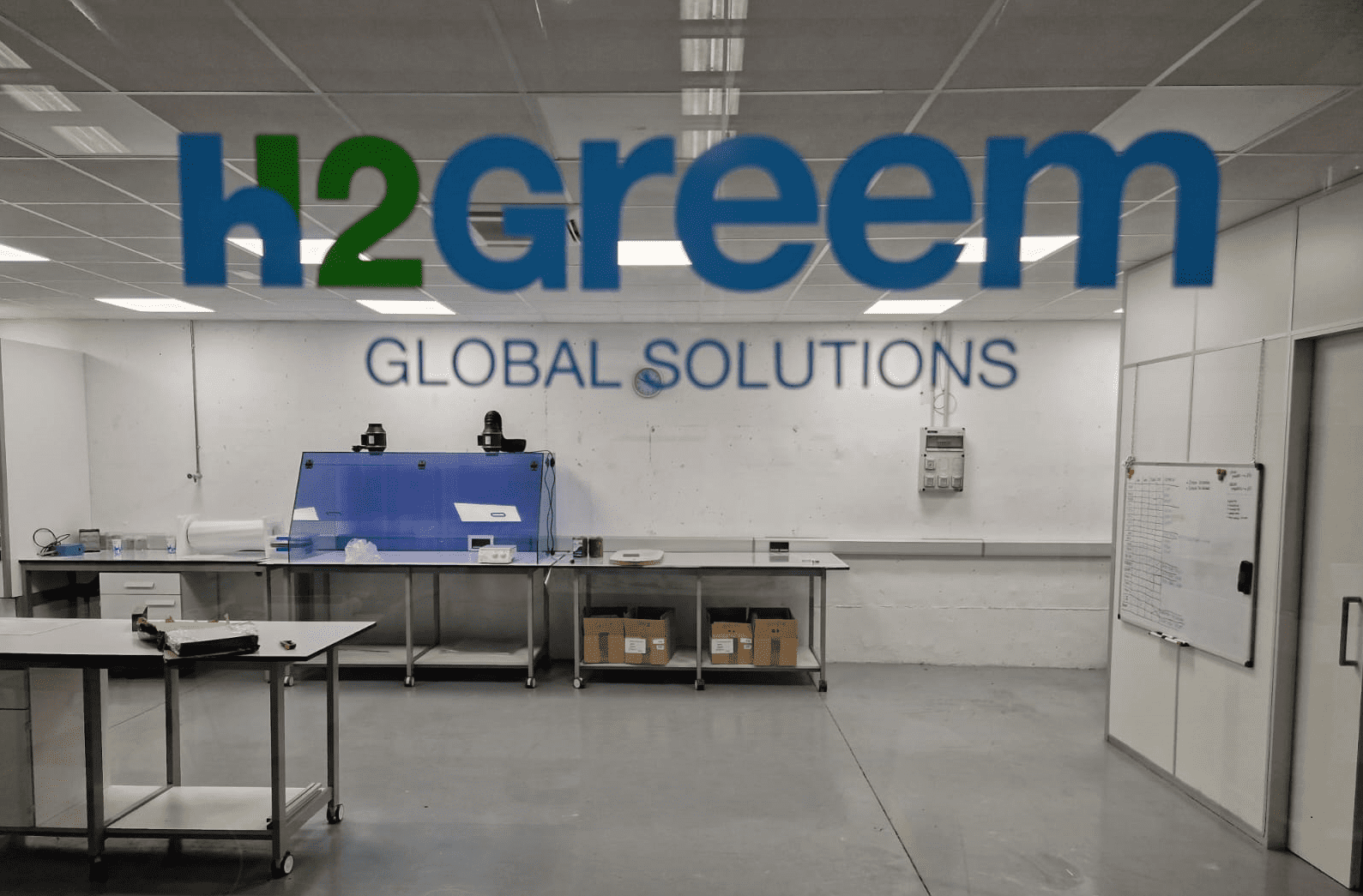 Parte del edificio laboratorio visto a través de un cristal que pone "H2Greem Gobal Solutions". Se ven elementos como mesas, con cajas debajo. Algunos elementos de laboratorio encima y una pizarra en una pared.